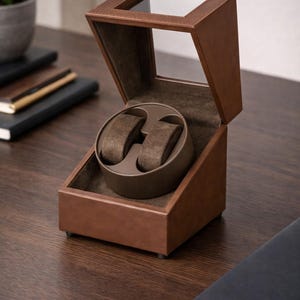 May include: A brown leather watch winder with a clear glass top. The interior has two watch holders with a soft, brown fabric. The winder sits on a dark wooden surface, suggesting a luxury item for watch enthusiasts.