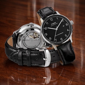 Pode incluir: Dois relógios automáticos Timex com pulseiras de couro preto e caixas prateadas. Um mostra as horas, o outro revela o mecanismo interno. O mostrador é preto com algarismos e ponteiros brancos. O texto "TIMEX AUTOMATIC THE WATERBURY" é visível.
