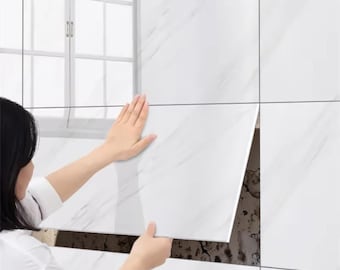 Puede incluir: Una persona instala baldosas blancas con diseño de mármol en una pared. Las baldosas rectangulares tienen vetas grises. La persona sostiene una baldosa, revelando la pared debajo. Una ventana es visible en el fondo.