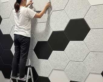 Puede incluir: Una persona en una escalera de mano instala paneles acústicos hexagonales en una pared. Los paneles están en un patrón de negro, blanco y gris. La persona lleva una camiseta blanca y pantalones negros.