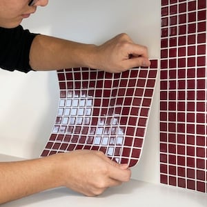 Könnte beinhalten: Ein flexibler Bogen aus tiefroten, quadratischen Fliesen mit weißen Fugen. Die Fliesenmatte hat eine glänzende Oberfläche und ein Gittermuster und ist für die Verwendung als Wandverkleidung oder Fliesenspiegel auf einer weißen Fläche vorgesehen.
