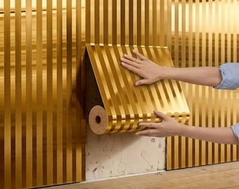Puede incluir: Papel pintado dorado con rayas verticales se aplica a una pared. El rollo de papel pintado está parcialmente desenrollado, revelando el patrón. La pared y el papel pintado tienen un acabado metálico similar, creando un aspecto cohesivo. El fondo es un suelo de madera.