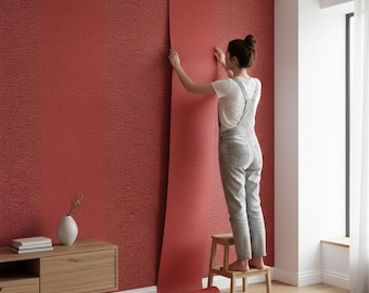 Puede incluir: Una persona está aplicando papel pintado rojo texturizado a una pared. El rollo de papel pintado está desenrollado, revelando un color sólido vibrante. La habitación cuenta con un mueble de madera, un jarrón y una ventana con cortina blanca. La pared es de color rojo intenso.