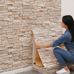 Pode incluir: Uma pessoa instalando painéis de parede de tijolo falso. Os painéis são uma mistura de bege, marrom e cinza, imitando a aparência de uma parede de pedra. Os painéis estão sendo aplicados em uma parede branca. A pessoa está usando uma camisa jeans e jeans.