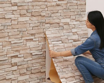 Könnte beinhalten: Eine Person installiert Wandpaneele in Ziegeloptik. Die Paneele sind eine Mischung aus Beige, Braun und Grau und ahmen das Aussehen einer Steinmauer nach. Die Paneele werden an einer weißen Wand angebracht. Die Person trägt ein Jeanshemd und Jeans.