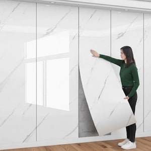 Puede incluir: Una mujer sostiene un panel grande blanco con un patrón de mármol contra una pared de paneles similares. Los paneles tienen un acabado brillante y están instalados en una pared blanca. La mujer lleva una camisa verde de manga larga y pantalones negros.