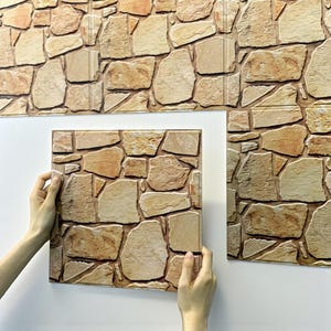 May include: Stone-effect wall panels in shades of tan, brown, and beige. The panels are designed to resemble natural stone. One panel is held up against a white wall, with other panels already installed.