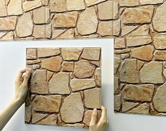 Könnte beinhalten: Steinoptik-Wandpaneele in Beige-, Braun- und Beigetönen. Die Paneele sind so gestaltet, dass sie das Aussehen von Naturstein nachahmen. Eine Platte wird vor einer weißen Wand gehalten, weitere Platten sind bereits installiert.