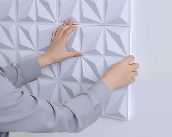 Puede incluir: Paneles de pared 3D geométricos blancos con un patrón triangular repetitivo. Los paneles se están instalando en una pared blanca. La imagen muestra las manos de una persona instalando los paneles.