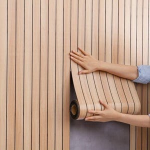 Può includere: Un rotolo di carta da parati color legno marrone chiaro con linee nere verticali viene applicato a una parete. La carta da parati viene srotolata da una persona. Anche la parete è rivestita con la stessa carta da parati.