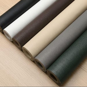 May include: A collection of seven rolled sheets of textured faux leather material in various colours including black, white, dark brown, beige, light grey, medium grey, and dark green, arranged in a diagonal row on a light wood surface.