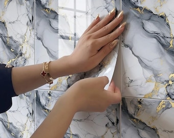 Puede incluir: Instalación de baldosas con diseño de mármol. Las baldosas presentan un diseño veteado blanco, gris y dorado. Se ven manos quitando el respaldo de la baldosa. Se ve una pulsera dorada con un dije de trébol rojo.