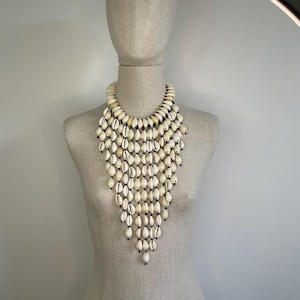 Puede incluir: Un collar llamativo compuesto por numerosas conchas marinas blancas y crema, cada una con una punta oscura. Las conchas están ensartadas para crear un diseño en cascada, estilo babero. El collar se exhibe en un maniquí.