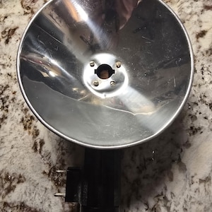 May include: A vintage silver-toned photographic flash reflector with a black base. The reflector has a circular, bowl-like shape with a central mounting point. The black base features electrical prongs.