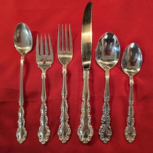 Op de afbeelding: Een set van zes zilverkleurige bestekstukken, waaronder lepels, vorken en een mes, gerangschikt op een rode ondergrond. Het zilverwerk heeft sierlijke, gedetailleerde handvatten. De lepels en vorken staan links, het mes in het midden.