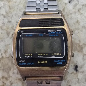 Puede incluir: Reloj digital vintage con caja dorada y correa de metal plateado. La esfera negra muestra la hora y las funciones. Funciones: alarma, hora y fecha. El reloj tiene forma rectangular.