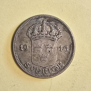 Könnte beinhalten: Eine kreisförmige, silberfarbene schwedische Münze aus dem Jahr 1916 mit einem zentralen Wappenschild mit drei Kronen, das von einer größeren Krone gekrönt wird. Der Text SVERIGE ist unten eingeprägt, die Jahreszahl 1916 ist durch das Schild getrennt.