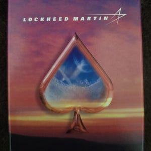Könnte beinhalten: Eine rechteckige Spielkartenschachtel mit einem Sonnenuntergangshimmel im Hintergrund in Violett-, Orange- und Gelbtönen. Der Text LOCKHEED MARTIN ist oben in Weiß aufgedruckt, mit einem großen Pik-Symbol in der Mitte, das einen blauen Himmel zeigt.