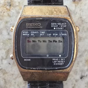 Puede incluir: Reloj digital Seiko Chronograph vintage con caja dorada y correa de cuero negro. La esfera muestra el día de la semana y varias etiquetas de funciones. El reloj tiene botones para iniciar/detener, contar y modo.