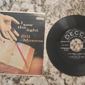 May include: Vintage vinyl record and album cover for Bill Monroe's "I Saw the Light." The album cover features hands reaching towards an open book. The record is black with a white and black label.