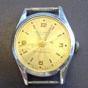 May include: A vintage wristwatch with a gold-coloured face and silver-coloured metal casing. The watch face has black numerals, hands, and the text "BICO SPECIAL" and "SWISS MADE". The watch face has a textured, patterned design.