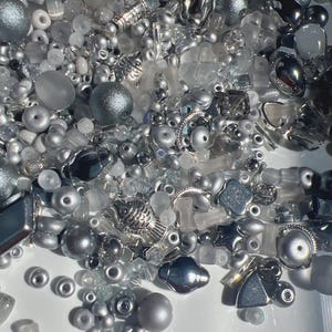 May include: A collection of assorted beads in various shades of silver, gray, and clear glass. The mix includes round, faceted, and textured beads, along with metallic charms shaped like fish and stars, all featuring a reflective finish.