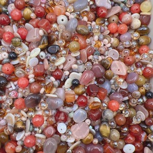 May include: A large collection of loose beads in various shapes, sizes, and textures. The assortment features a warm color palette dominated by shades of red, orange, brown, cream, and grey, including smooth, faceted, and irregular stone-like beads.