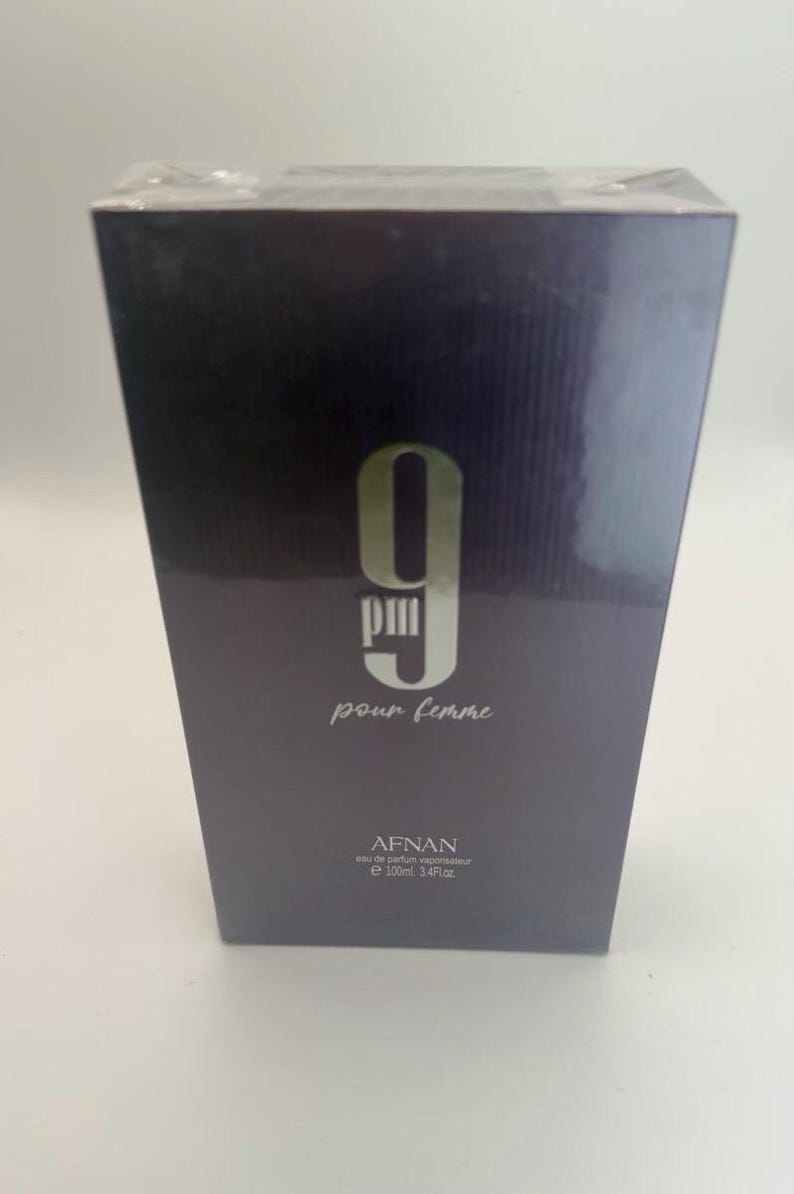 Puede incluir: Caja de perfume gris oscuro con el texto "9 PM pour femme" en blanco. La marca "AFNAN" est&aacute; impresa debajo, junto con el volumen: 100 ml, 3,4 fl oz.