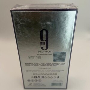 Puede incluir: Caja plateada de eau de parfum "9 pm pour femme". La caja presenta el n&uacute;mero "9" en negrita, con las palabras "pour femme" debajo. La caja tambi&eacute;n incluye los ingredientes y otros detalles.