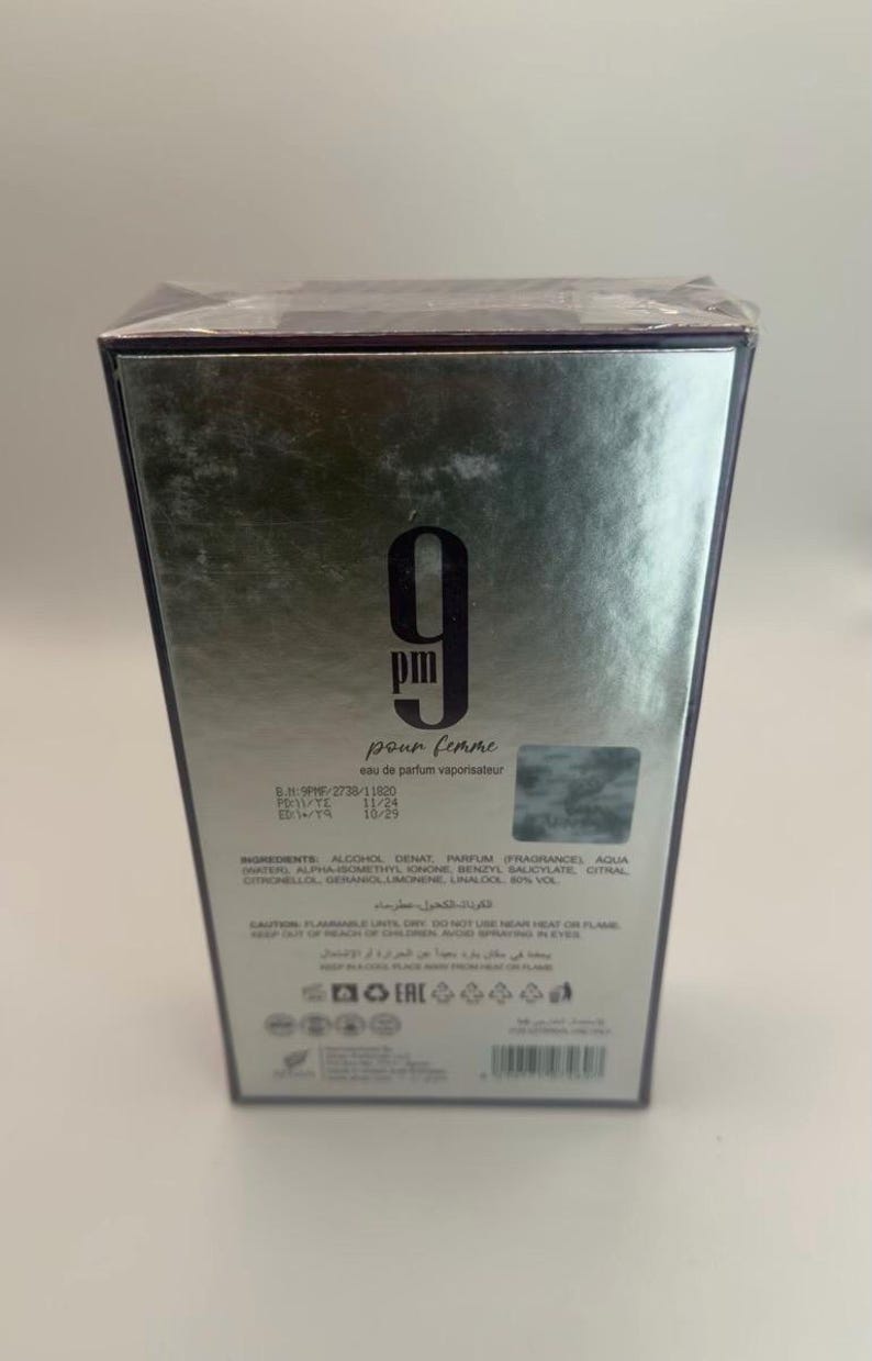 Puede incluir: Caja plateada de eau de parfum "9 pm pour femme". La caja presenta el n&uacute;mero "9" en negrita, con las palabras "pour femme" debajo. La caja tambi&eacute;n incluye los ingredientes y otros detalles.