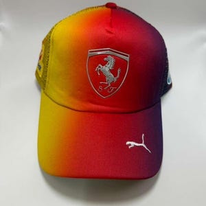 May include: A gradient-colored baseball cap with a yellow to red fade. The cap features the Ferrari logo on the front and a Puma logo on the brim. The mesh back is yellow.