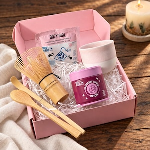 Peut inclure: Une boîte cadeau rose contenant un fouet à matcha en bambou, une cuillère en bambou, une tasse en céramique rose et blanche, une boîte de Raspberry Ripple Matcha et un sachet de tisane Dozy Girl, le tout sur un lit de papier déchiqueté blanc.