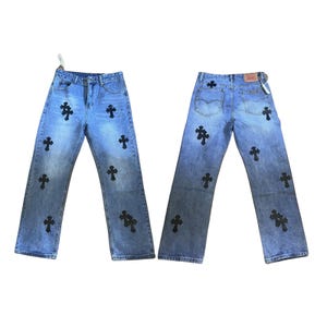 May include: Light blue denim jeans with black cross appliques. The jeans have a classic five-pocket design and a straight leg fit. The back of the jeans shows a Levi's tag. The jeans are a medium wash and have a button and zip closure.