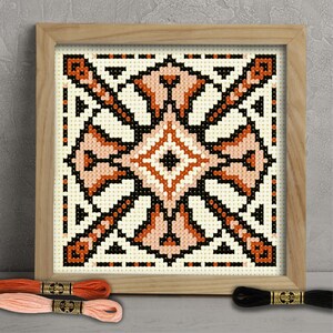 Peut inclure: Une œuvre d'art au point de croix encadrée avec un motif géométrique en crème, noir et nuances d'orange. L'œuvre est dans un cadre en bois clair. Des échevettes de fil à broder orange et rose sont au premier plan, avec une échevette noire.