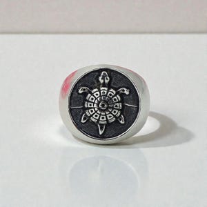 Puede incluir: Anillo de plata con un diseño detallado de tortuga. La tortuga está centrada sobre un fondo negro en la cara del anillo. El anillo tiene una superficie pulida y reflectante.