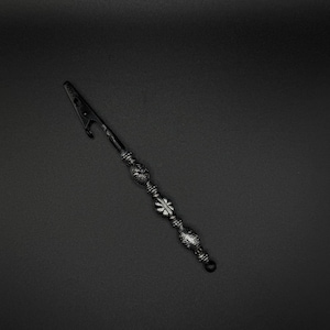 May include: A black metal roach clip featuring a pointed, angled tip and a handle decorated with three distinct silver-toned beads. The beads have intricate, embossed floral and geometric patterns, providing a textured grip along the slender shaft.