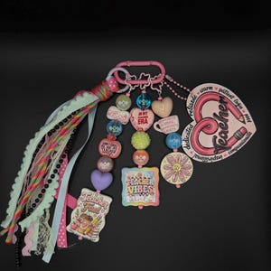 May include: A pink carabiner keychain featuring a collection of colorful beads, charms, and fabric ribbons. The charms include text such as Teacher Fuel, In My Teacher Era, Teacher Vibes, and a heart-shaped tag with the word Teacher and descriptive adjectives.