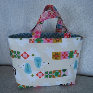 May include: A small fabric tote bag featuring a repeating pattern of white dachshund dogs with blue ears and colorful geometric quilt-style blocks on their bodies. The bag has two matching handles with a pink and multi-colored geometric pattern.