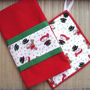 Puede incluir: Un juego de paño de cocina y agarradera rojos. La toalla tiene una banda blanca con estampado de muñeco de nieve y ribete verde. La agarradera tiene un estampado de muñeco de nieve a juego con ribete rojo y un lazo para colgar.