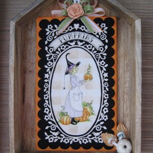 May include: Wooden framed Halloween decoration featuring a witch holding a pumpkin. The artwork is set against an orange background with a black and white scalloped border. The word "PUMPKINS" is displayed on a banner.
