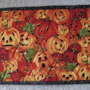 May include: A rectangular Halloween-themed quilted table runner. The fabric features a repeating pattern of orange and red jack-o'-lanterns with various expressions, interspersed with green leaves. The runner is bordered with a dark gray fabric.