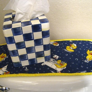 Puede incluir: Un dispensador de pañuelos a cuadros azules y blancos se encuentra sobre una funda de asiento de inodoro azul marino con un borde amarillo. La funda presenta un patrón de patitos de goma amarillos en bañeras y burbujas.