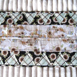 May include: A quilted, hexagonal table runner featuring a central panel with a pumpkin and floral pattern. The edges are adorned with alternating black sunflower and plaid triangle patterns. The color palette includes white, black, brown, and shades of green.