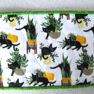May include: A rectangular quilted fabric with a white background featuring a repeating pattern of black cats and potted plants. The border is green. The cats are in various poses, and the plants are in yellow and brown pots.