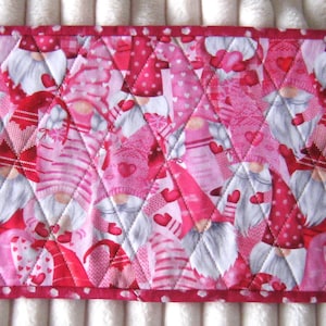May include: A rectangular quilted fabric featuring a repeating pattern of gnomes in shades of pink, red, and white. The gnomes have white beards and red hats, with heart accents. The border is a red polka dot pattern.