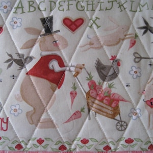 May include: Quilted fabric with a whimsical bunny theme. Features a bunny in a top hat pushing a cart of carrots, along with other bunnies, chickens, and floral and heart motifs. The word "Bunny" is printed in red.