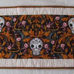 May include: A decorative table runner featuring a repeating pattern of sugar skulls, flowers, and foliage in orange, black, and pink. The central design is framed by a white border with a quilted pattern and a brown trim.