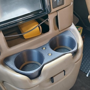 May include: A custom-fit, black dual cup holder insert for a vehicle's centre console. The insert replaces the original console components and features two circular cup holders with textured bases. The surrounding console is tan.