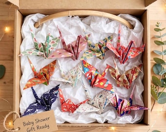 May include: A cardboard box containing twelve folded paper cranes in various colorful floral and geometric patterns. The cranes are suspended from a curved wooden arch. A rectangular tan tag attached to the box reads Baby Shower Gift Ready.