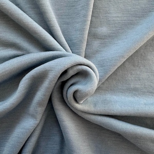 Może przedstawiać: Zbliżenie jasnoniebieskiej tkaniny o miękkim, teksturowanym wyglądzie. Tkanina jest udrapowana i zakręcona, tworząc efekt spirali. Materiał wydaje się być dzianiną lub tkaniną, z widocznymi nićmi i subtelnym połyskiem. Całość prezentuje się gładko i zachęcająco.